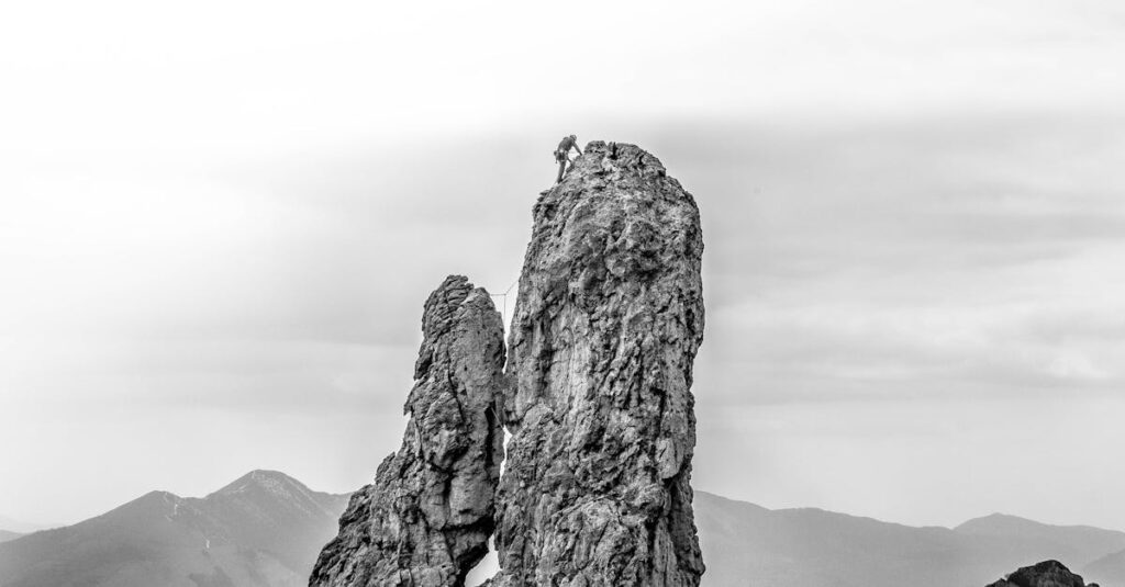Adventurous climber reaching the summit of a towering rocky mountain, set against a moody sky.