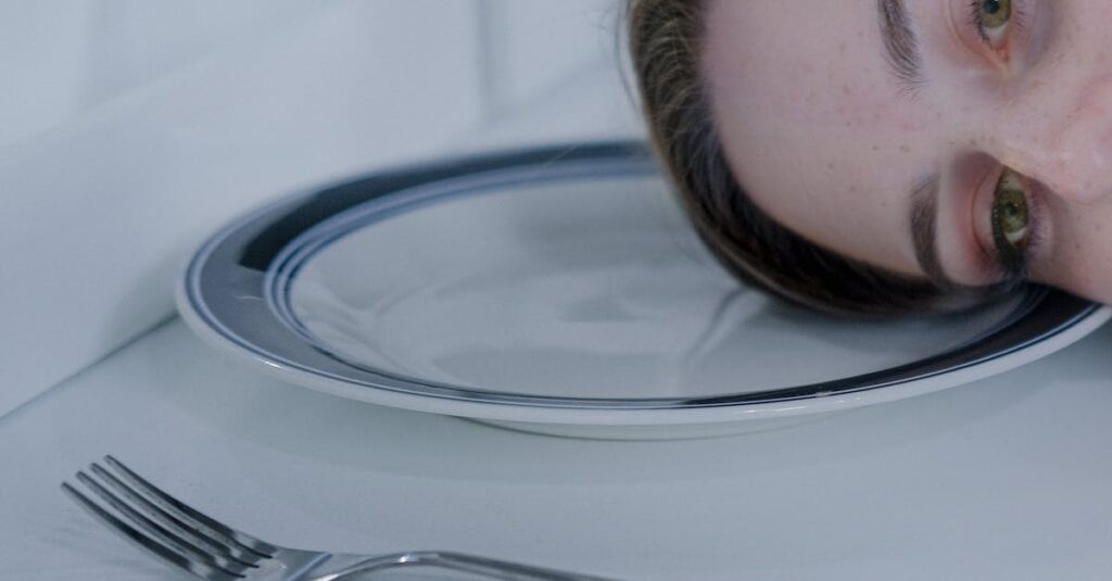 A woman rests her head on a plate next to a fork, exuding a contemplative mood.