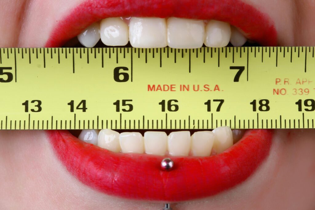 Close-up of red-lipsticked mouth holding a yellow tape measure horizontally, demonstrating measurement concept.
