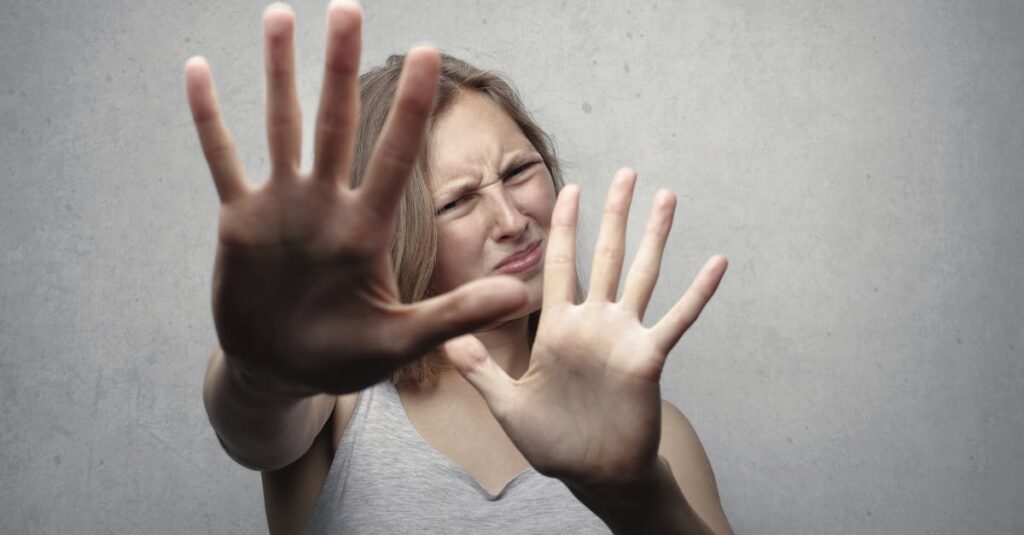 Portrait of a fearful woman in a gray tank top with hands pushed forward against a gray background.