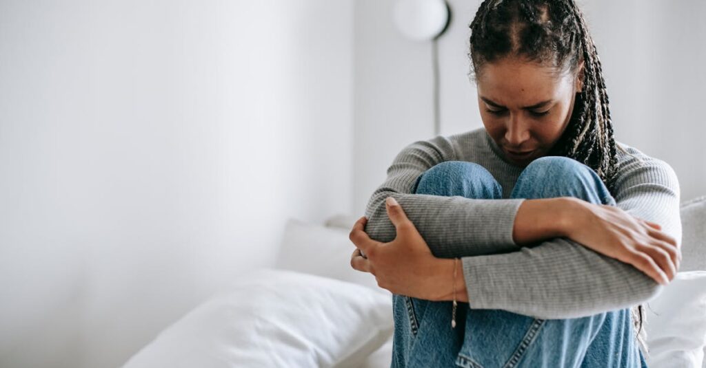 A young woman embraces her knees while sitting on a bed, appearing thoughtful and introspective.