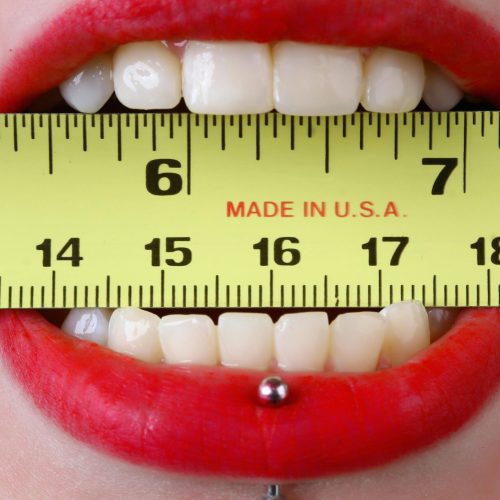 Close-up of red-lipsticked mouth holding a yellow tape measure horizontally, demonstrating measurement concept.