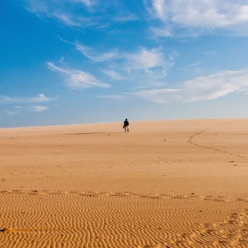 A lone traveler walks across a vast and serene desert under a bright blue sky.